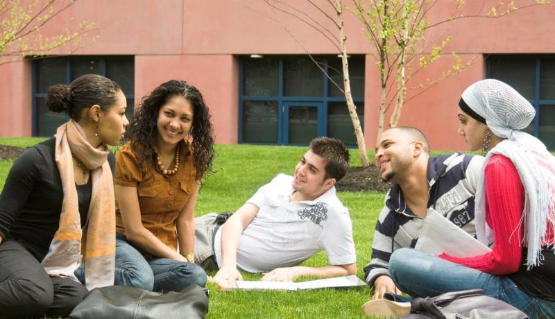 A diverse group of 3 female and 2 male students sitting and talking on a lawn on campus.