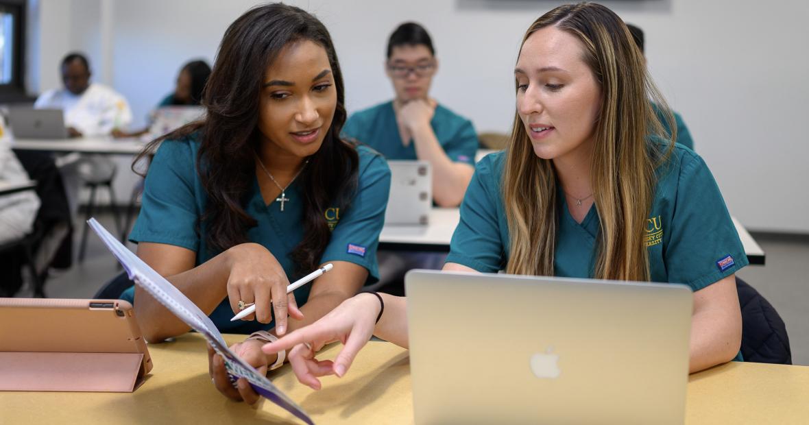 Two nursing students examine study materials while using a macbook and iPad