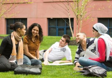 A diverse group of 3 female and 2 male students sitting and talking on a lawn on campus.