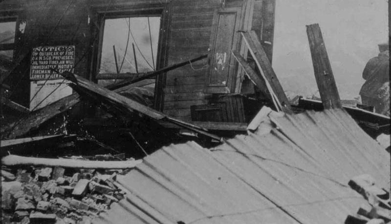 Black and white photograph from the aftermath of the Black Tom explosion of one wall standing surrounded by bricks and wooden debris