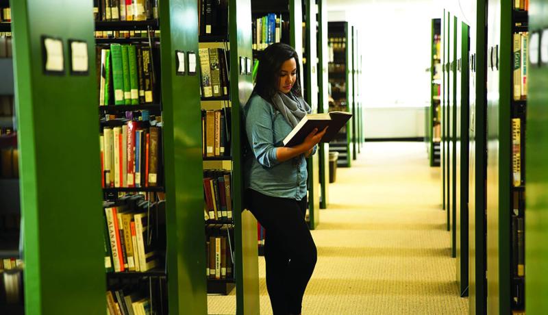 Student reading a large book in the library next to aisles of books.