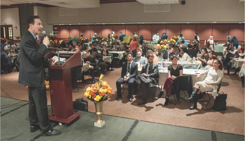 Attendees of the Open Arms, Open Hearts, Open Doors event site in the audience while a man speaks at the podium on stage.