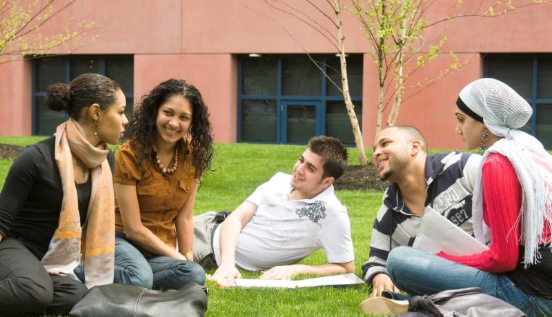 A diverse group of 3 female and 2 male students sitting and talking on a lawn on campus.