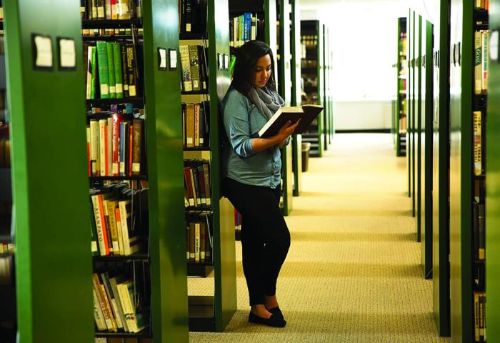 Student reading a large book in the library next to aisles of books.