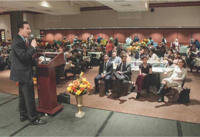 Attendees of the Open Arms, Open Hearts, Open Doors event site in the audience while a man speaks at the podium on stage.
