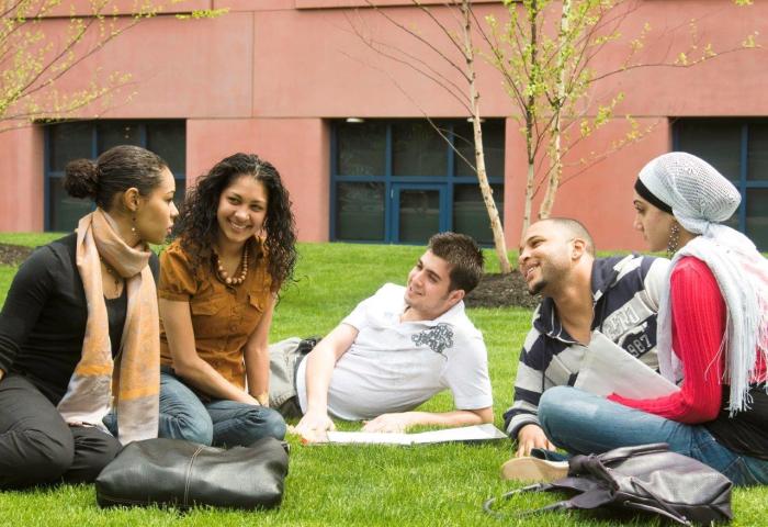 A diverse group of 3 female and 2 male students sitting and talking on a lawn on campus.