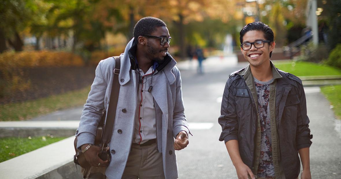 Two male students walking and talking through campus Fall background