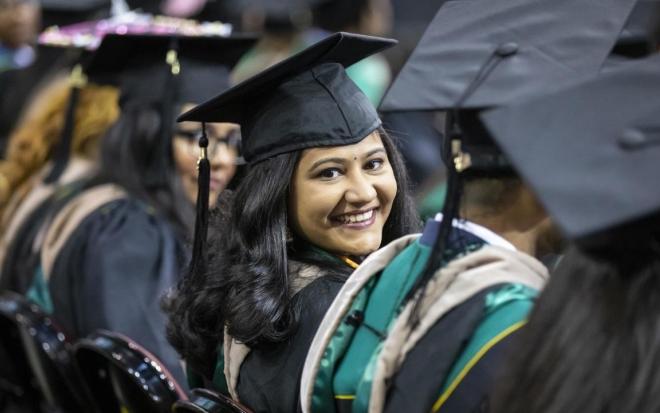 Woman in cap and gown sitting in row at commencement ceremony smiling