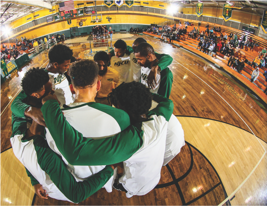 NJCU men's basketball team huddled on the court.