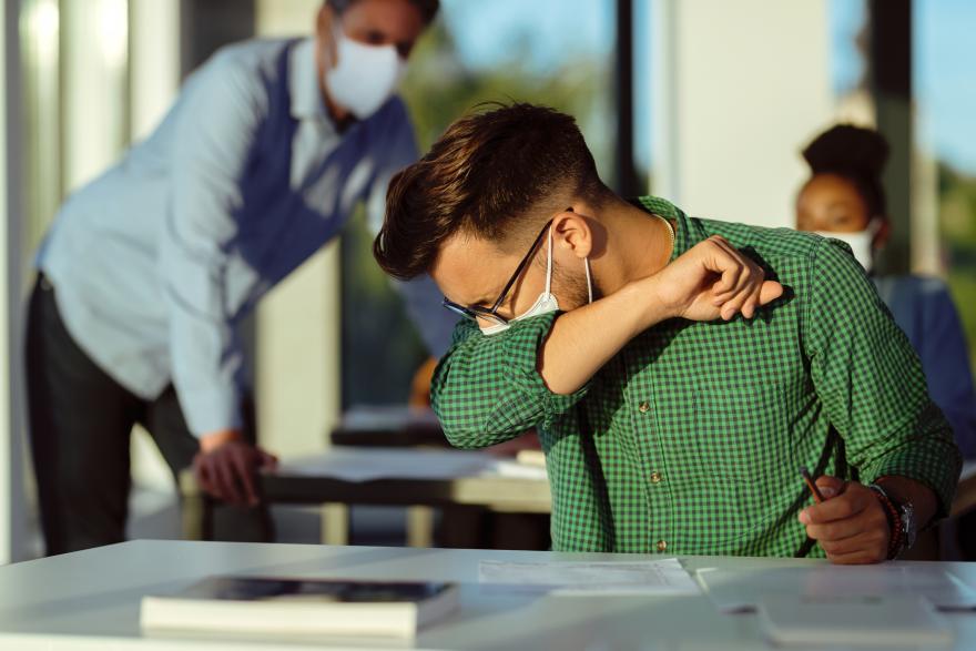 Young man sneezing into elbow in classroom with people in the background