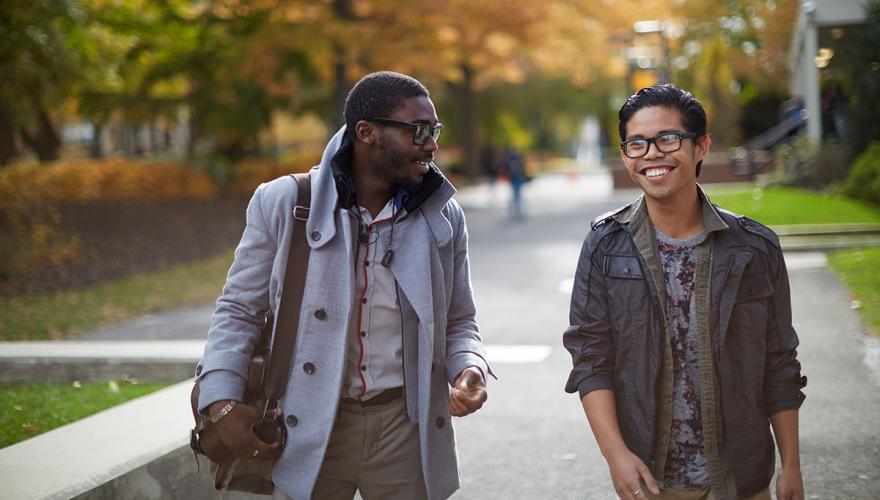 Two male students walking and talking through campus Fall background