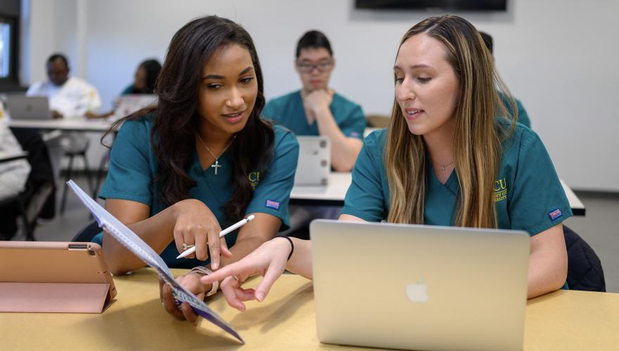 Two nursing students examine study materials while using a macbook and iPad