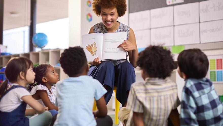Female Teacher Reads To Multi-Cultural Elementary School Pupils Sitting On Floor In Class