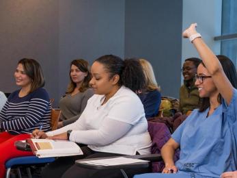 Classroom with students, one of whom is in scrubs and raising her hand