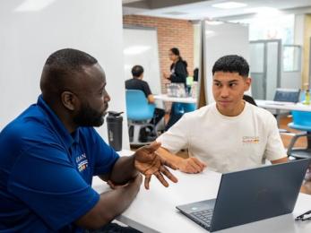 A man is providing technical support to a student with a laptop on the desk.